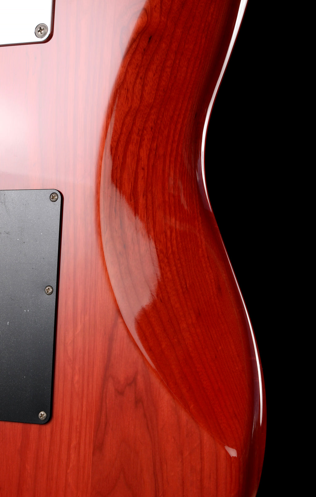 Fender Japan Michiya Haruhata Stratocaster Transparent Pink 2021: Close-up of a glossy reddish-brown wooden guitar body with visible wood grain, a black pickguard, and part of a white control plate against a black background.
