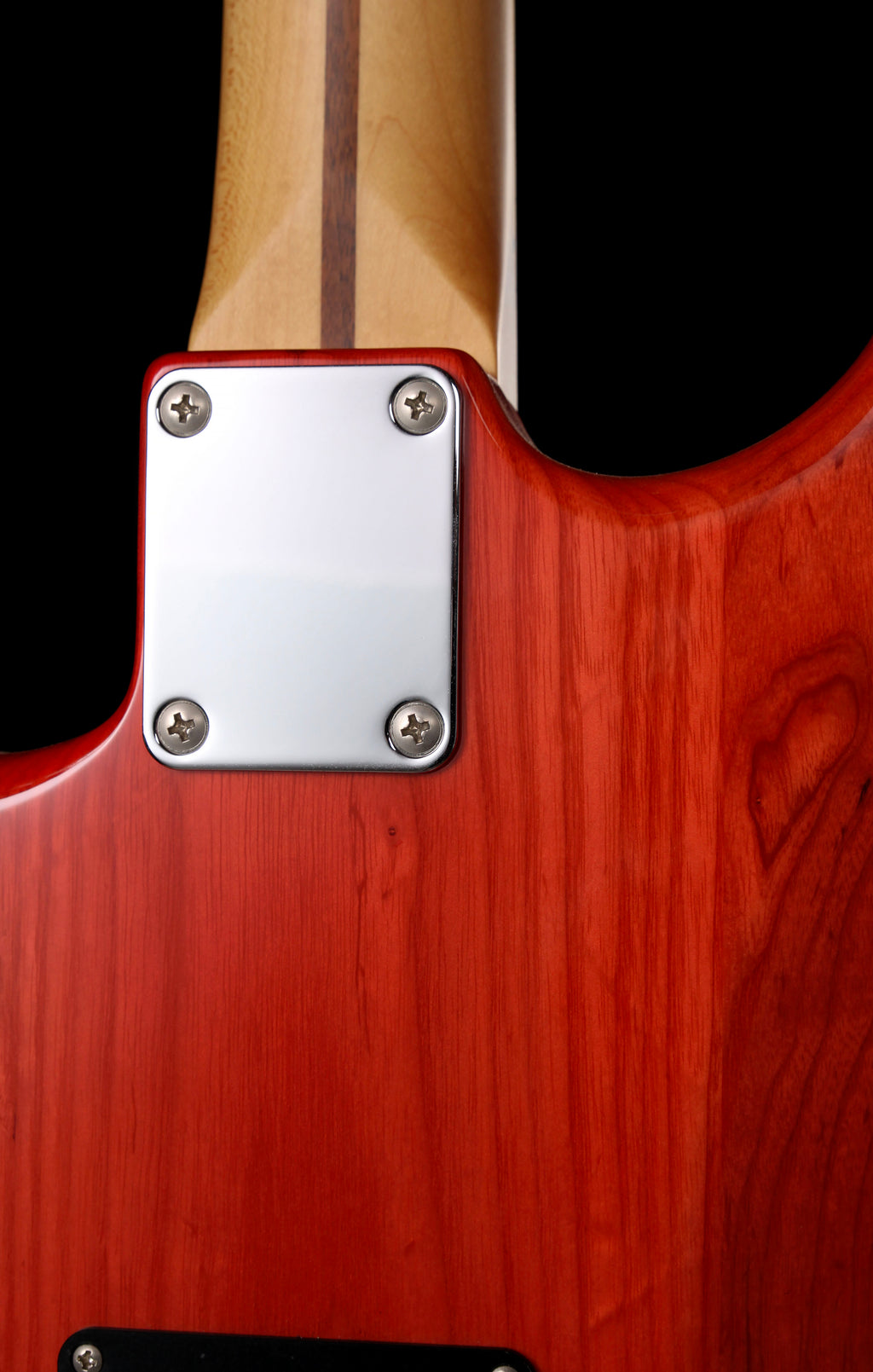 Fender Japan Michiya Haruhata Stratocaster Transparent Pink 2021: Close-up of the back of an electric guitar showing a glossy reddish-brown wood body, a silver metal plate secured by four screws, and part of a light wood neck against a black background.