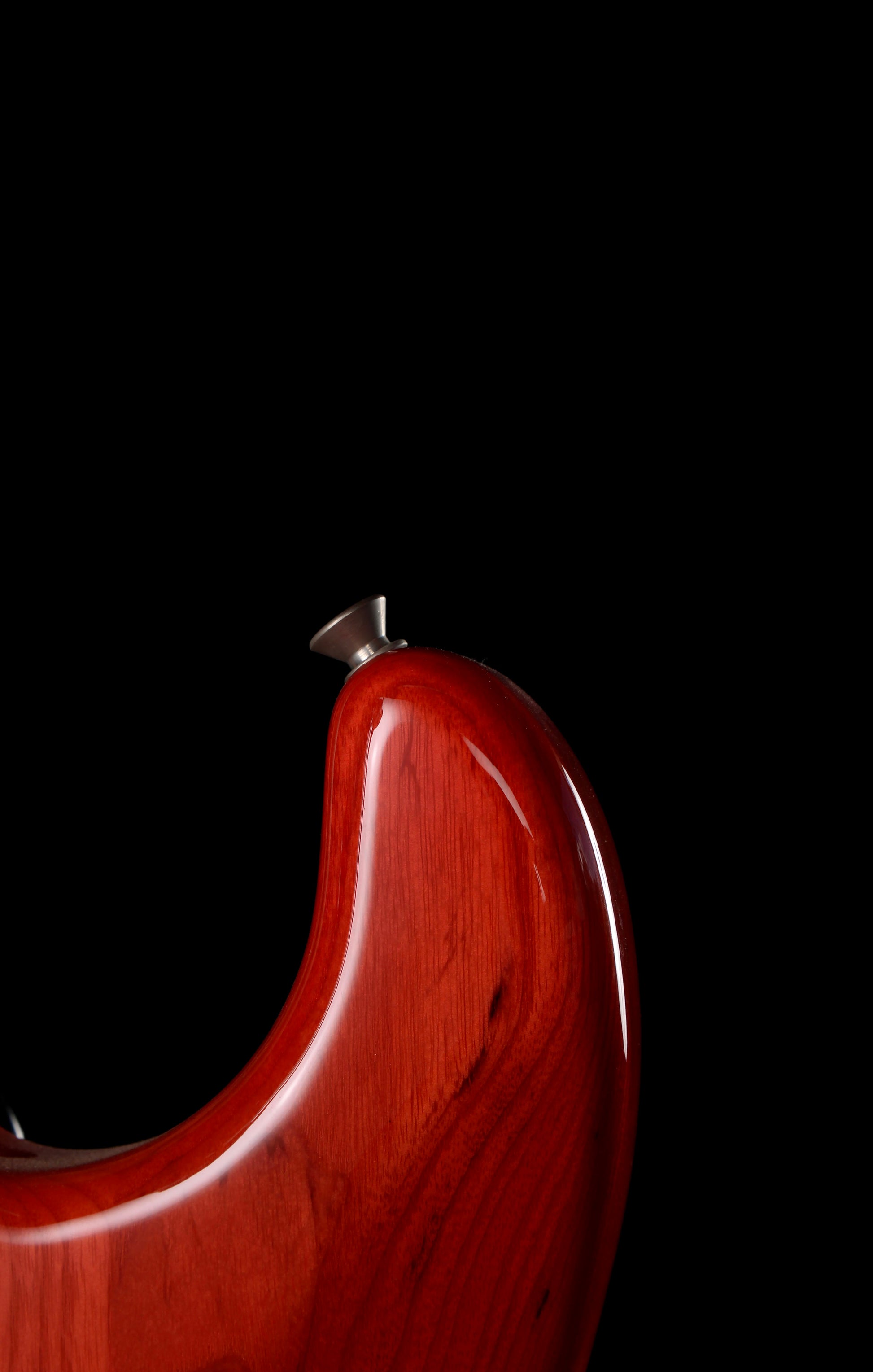 Fender Japan Michiya Haruhata Stratocaster Transparent Pink 2021: Close-up of a glossy reddish-brown wooden guitar body corner with a silver tuning peg against a solid black background.