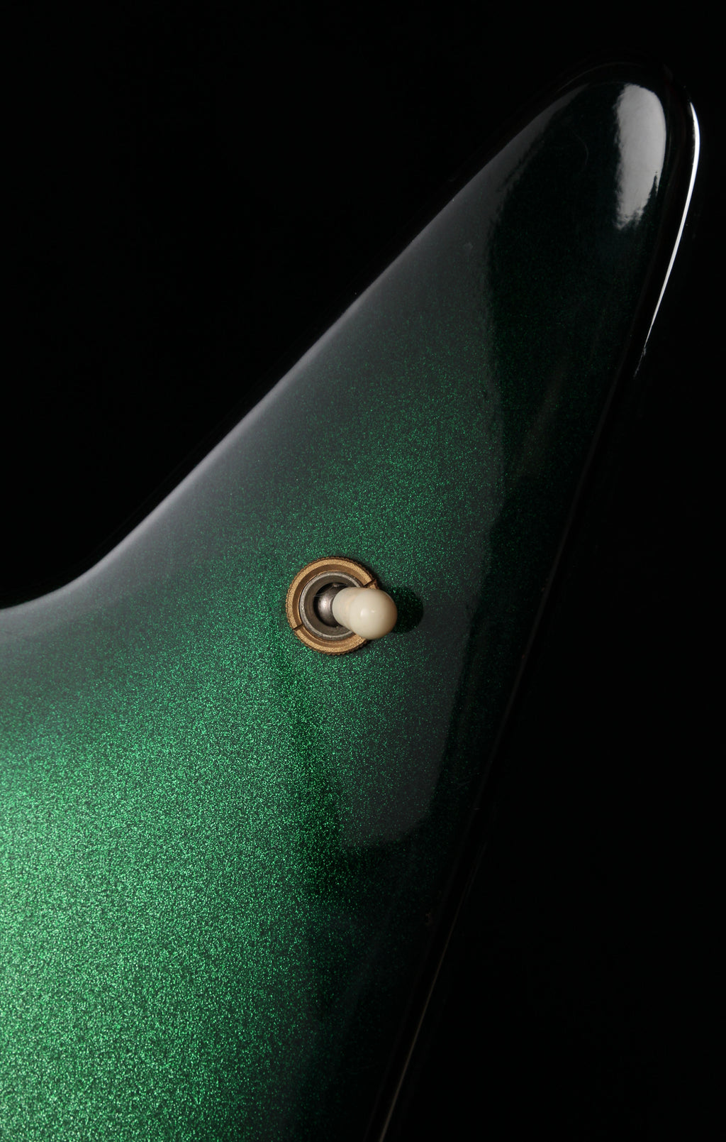 Fernandes EX Explorer Special Order SGS Sparkle Green Sunburst MIJ 1984: Close-up of a glossy dark green guitar body with a small gold and white control switch, set against a solid black background.